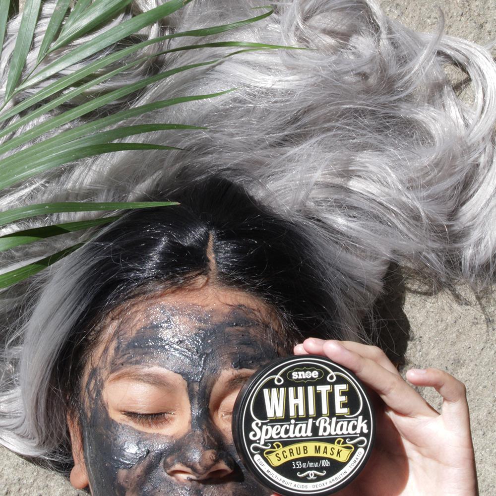 A woman holding a SNOE WHITE SPECIAL BLACK Bleaching Whitening Facial Scrub Mask with a palm tree in the background.