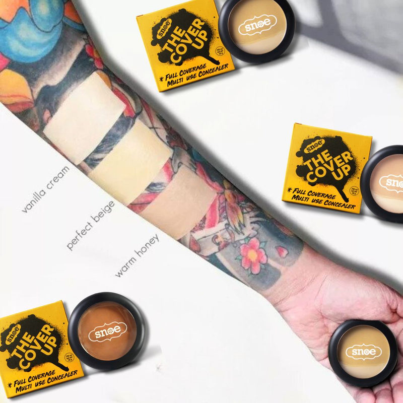 A tattooed arm is elegantly displayed with swatches of concealer skillfully hiding the ink, showcasing its transformative beauty. Three shades—vanilla cream, perfect beige, and warm honey—exhibit the make-up's coverage prowess. Resting nearby are three yellow-packaged concealers titled "Buildable Concealer in PERFECT BEIGE" from THE COVER UP brand.