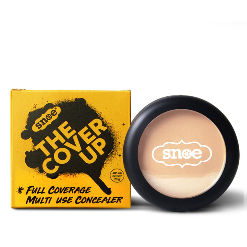 A round container of concealer is displayed next to its yellow box packaging, embodying elegance in beauty. The box features bold black text reading "THE COVER UP," with a design resembling a make-up brush sweep. The concealer, named Buildable Concealer in VANILLA CREAM, is housed in a circular case that also bears the Snoe logo on its transparent lid.
