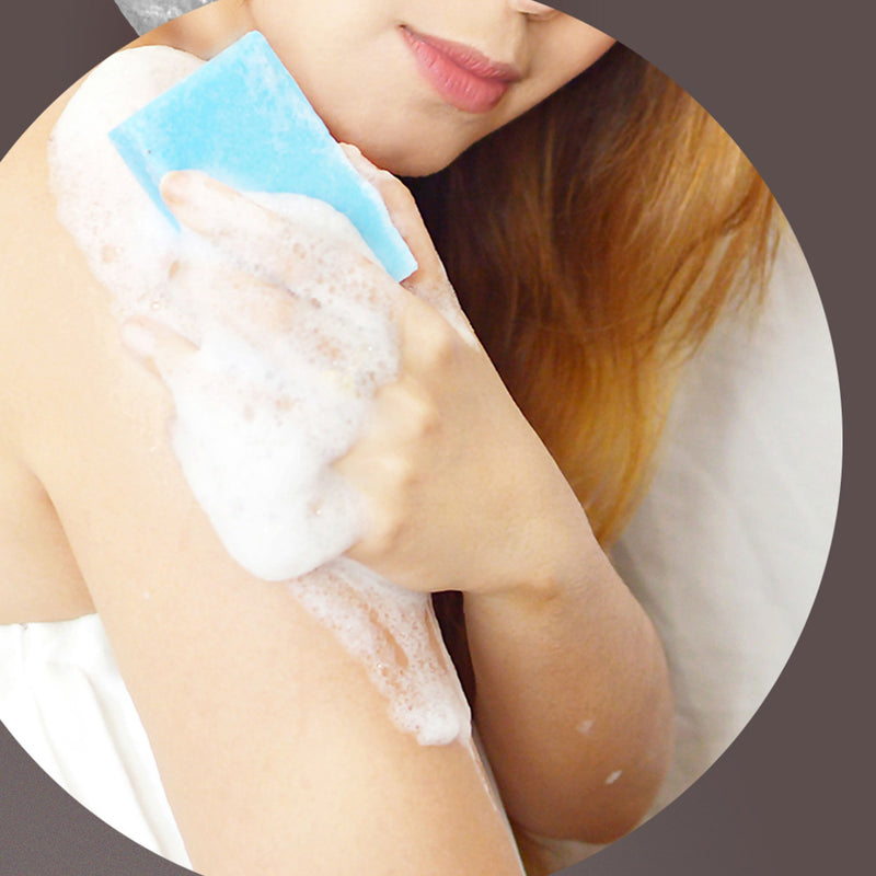 With shoulder-length hair, a person enjoys a beauty ritual using S-SKIN Naturals' EPSOM SALT: Magnesium Sulfate Body Soap & Scrub, a blue bar of soap. Their hand and upper arm are enveloped in white, frothy suds. Draped in a white towel, only their upper torso and part of their face appear against the neutral background.