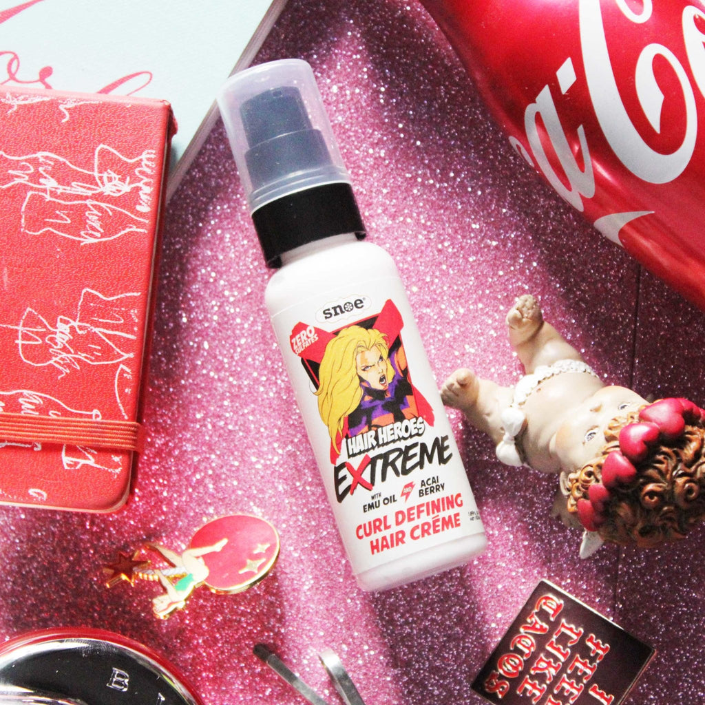 A bottle of HAIR HEROES EXTREME Curl Defining Hair Crème and other items on a pink table, featuring beauty products.