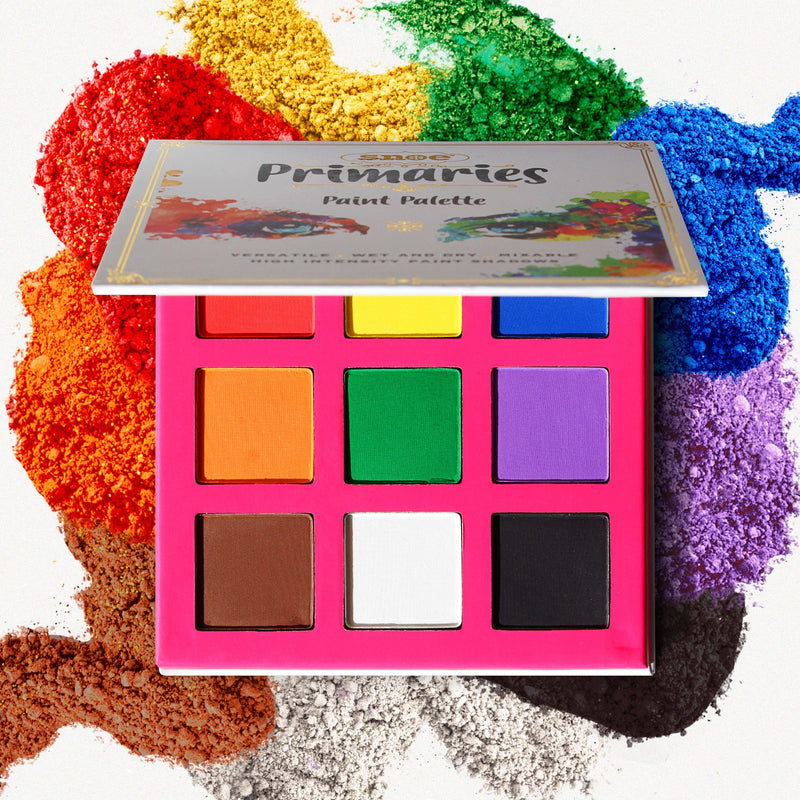 The PAINT EYESHADOW PALETTE by PRIMARIES is showcased, featuring a vibrant and versatile range of high-intensity shades including yellow, green, red, orange, blue, purple, brown, and black. With its lid open to unveil the stunning colors inside, splashes of matching hues are artistically arranged around the palette.