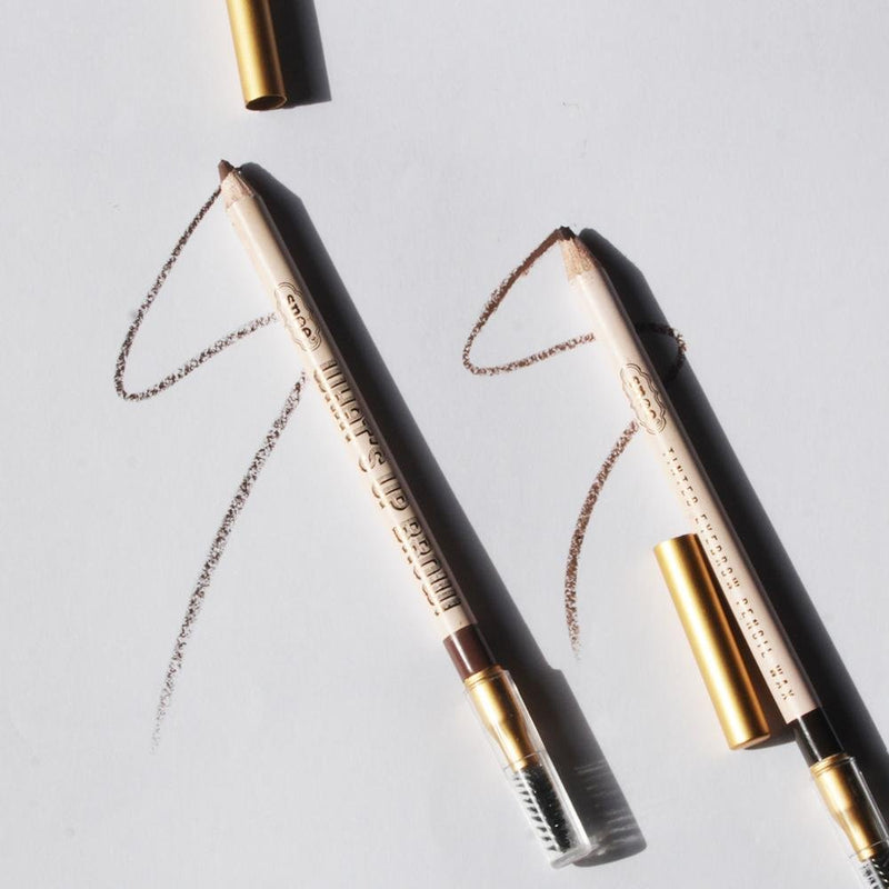 Two Tinted Eyebrow Pencil Wax items in KHAKI from WHATS UP BROW! with gold caps are placed diagonally on a white surface, capturing the essence of beauty. Each pencil features a brown line drawn from its tip, curving gracefully in an arch. These essential makeup tools are equipped with spoolie brushes, casting delicate shadows.