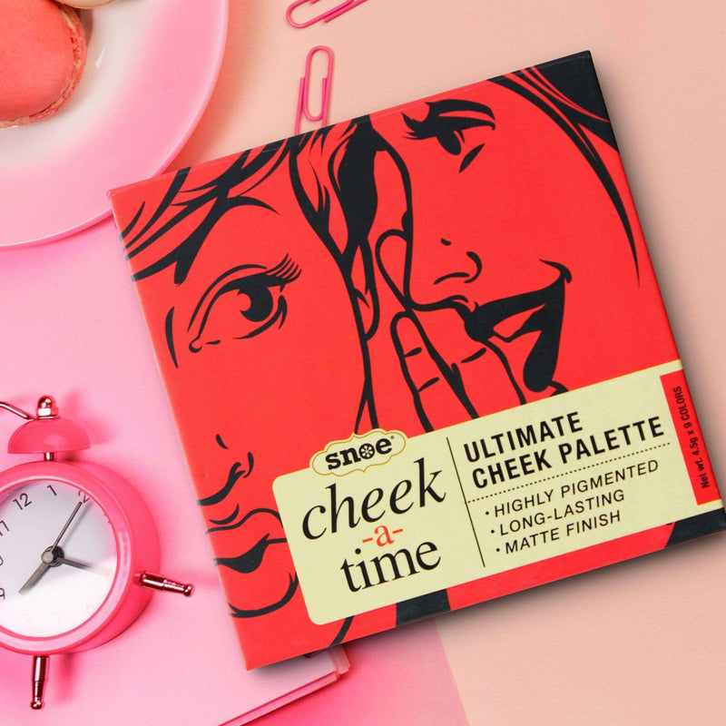 An eye-catching cheek palette, "Ultimate Cheek Palette" by CHEEK A TIME, is prominently displayed with its bold design featuring illustrated faces on a vibrant red backdrop. Ideal for beauty enthusiasts, the packaging emphasizes its high pigmentation, long-lasting effect, and matte finish. Surrounding it are a pink alarm clock, paper clips, and a macaron.